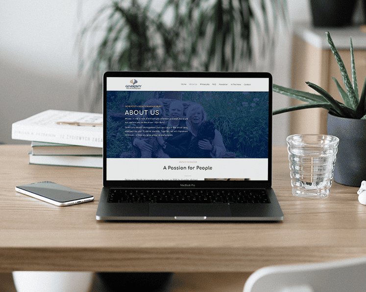 iMc-GWM MacBook mobile Laptop on a wood desk displaying the “About Us” page of Generosity Wealth Management’s website, with books, a phone, and glass nearby.