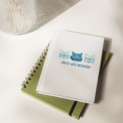 A white hardcover journal featuring three illustrated teal cats wearing glasses and the phrase “Create with Intention” on the cover, resting on top of a green notebook under soft natural light.